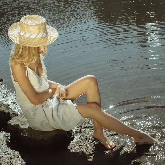 Phoebe Dynevor sitting beside the water in a white dress and straw hat.