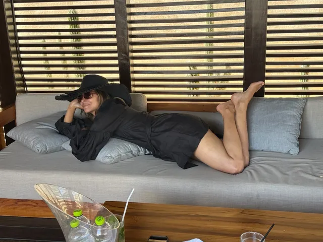 Halle Berry reclining barefoot on a sofa in a dark outfit and cap.