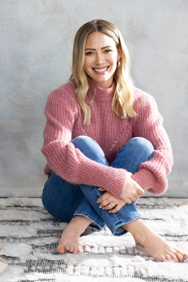 Hilary Duff sitting barefoot on steps in a pink sweater and jeans.