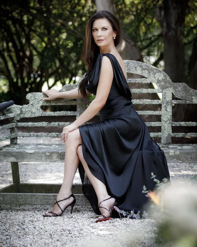 Catherine Zeta-Jones seated on an outdoor bench in a black dress and heels.