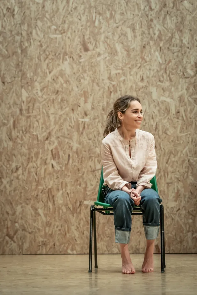 Emilia Clarke sitting barefoot in a cream blouse and rolled jeans.
