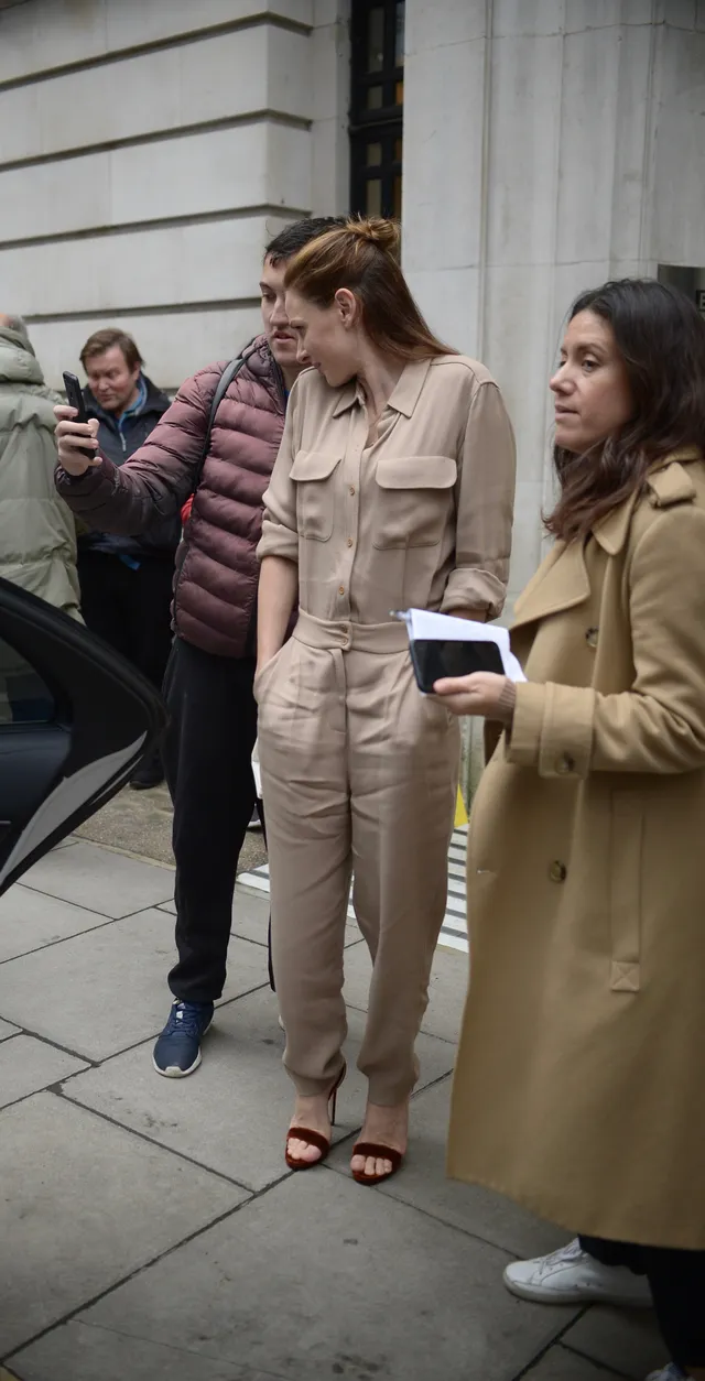 Rebecca Ferguson standing outdoors in a beige jumpsuit and open-toe heels beside companions.