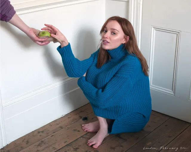 Phoebe Dynevor seated on a wooden floor in a blue sweater.