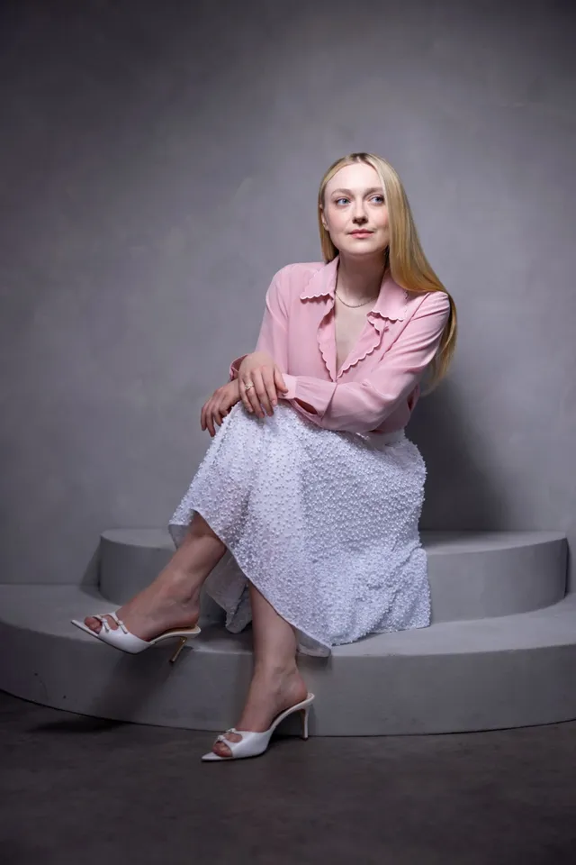 Dakota Fanning sitting in a pink jacket, patterned skirt and silver heels.