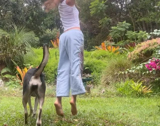 Freya Allan jumping barefoot on grass beside a dog.