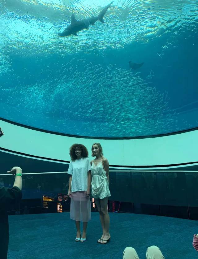 Phoebe Dynevor posing beside another woman at an aquarium in casual shoes.