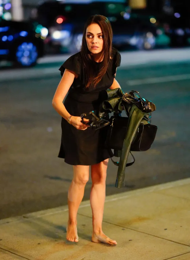 Mila Kunis walking barefoot on a pavement while carrying shoes.