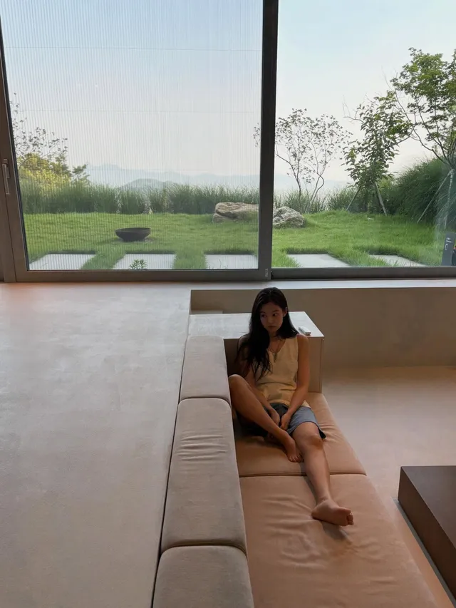 Jennie Kim sitting barefoot on an indoor lounger beside a garden window.