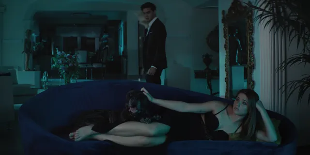 Nicole Wallace reclining barefoot on a sofa in a film scene.