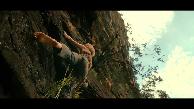Charlize Theron a screen scene climbing on a rock face barefoot.