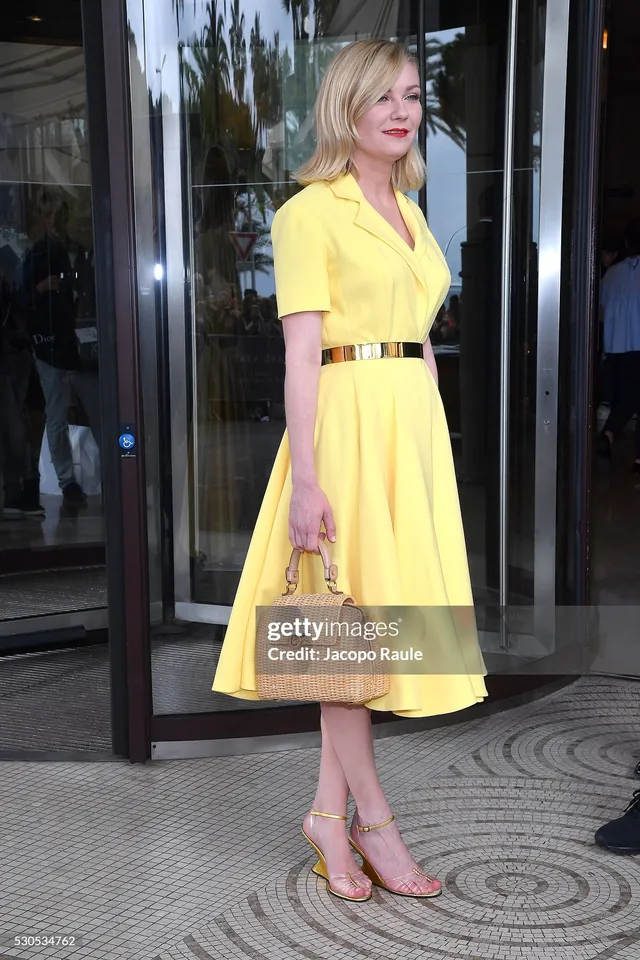 Kirsten Dunst standing in a yellow dress and wedge heels near an entrance.