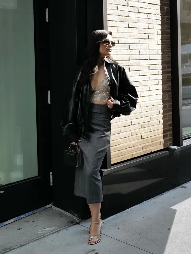 Rachel Zegler standing outside in a black jacket, grey skirt and heeled sandals.