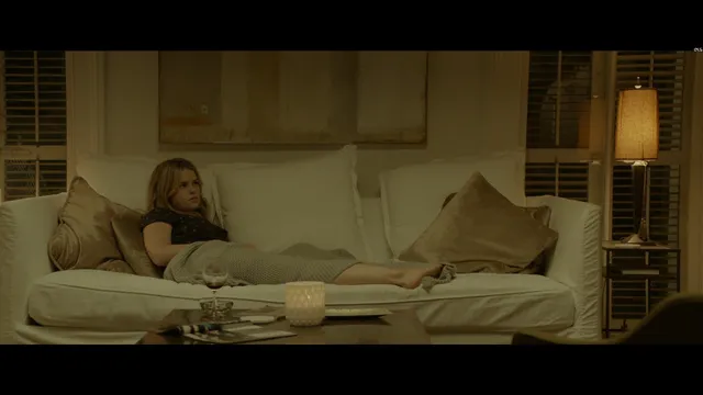 Alice Eve reclining on a pale sofa in a screen scene.