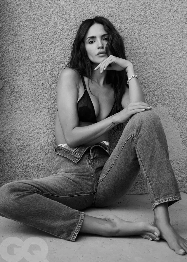 Eiza González sitting barefoot in jeans for a monochrome portrait.