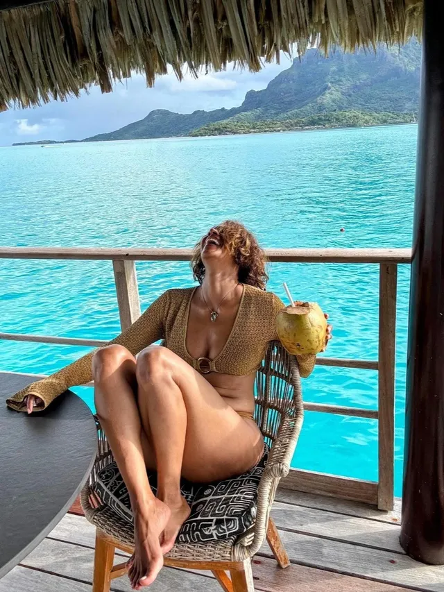 Halle Berry sitting barefoot on a chair beside turquoise water.