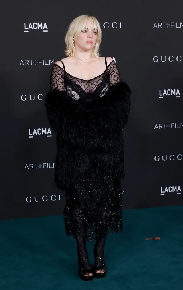Billie Eilish posing against an event backdrop in a lace dress and heels.