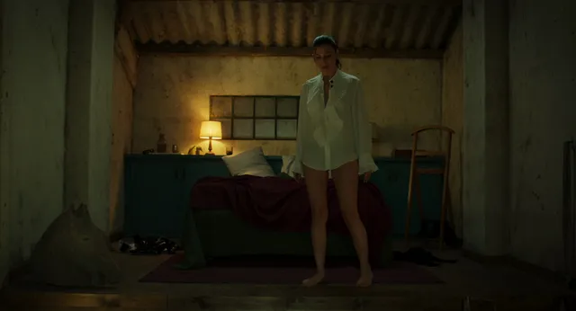 Úrsula Corberó standing barefoot in a pale shirt and shorts in a bedroom scene.