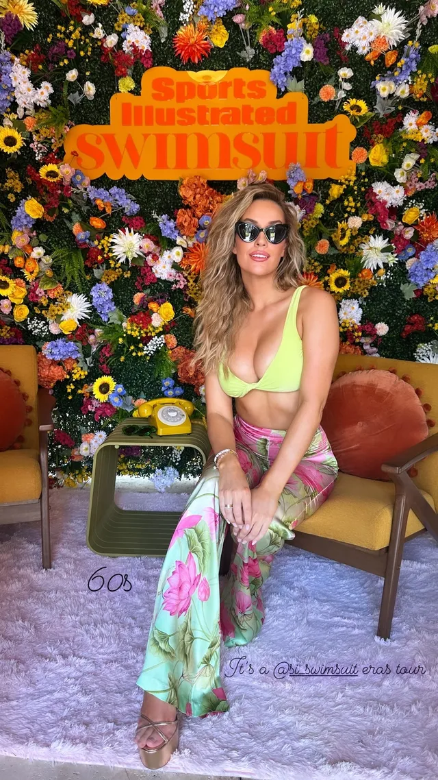 Paige Spiranac wearing heeled sandals with bright floral trousers and a green top at an event.