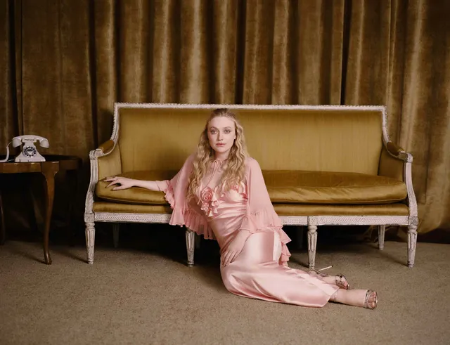 Dakota Fanning sitting on the floor beside a sofa in a pink dress and heels.