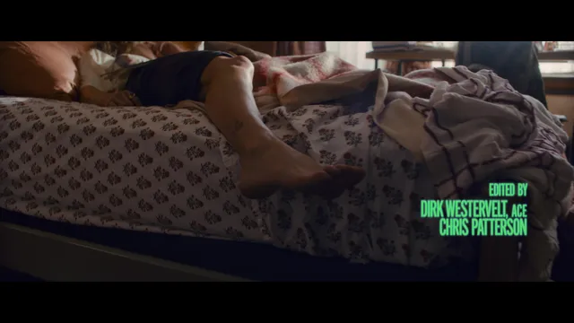 Samara Weaving resting a bare foot over the edge of a bed in a film scene.