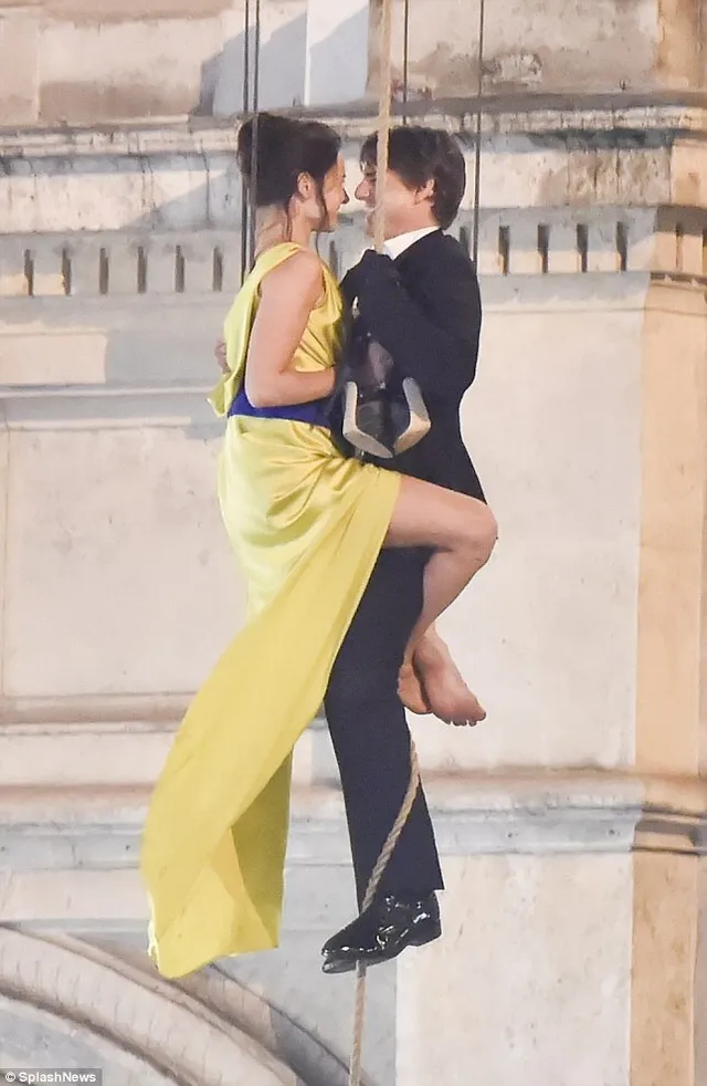 Rebecca Ferguson posing barefoot in a yellow dress beside a companion on a rooftop.