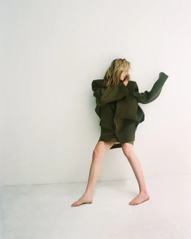 Kiernan Shipka standing barefoot in a dark jacket for a studio portrait.