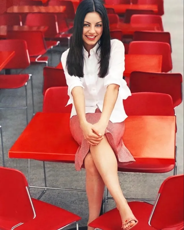 Mila Kunis sitting among red chairs in a pink skirt and heels.
