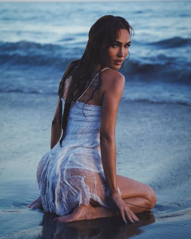 Megan Fox kneeling beside the sea in a white dress.