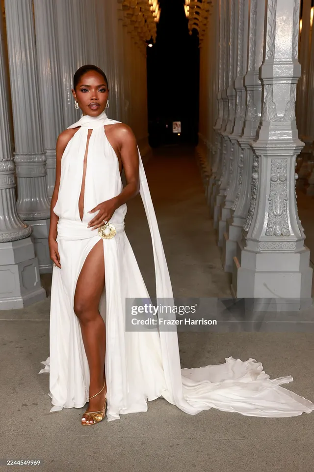 Ryan Destiny posing in a white halter gown and gold heels at an event.