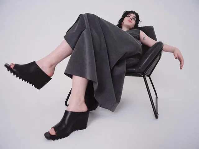 Sophie Thatcher reclining in a studio chair in wide trousers and platform heels.