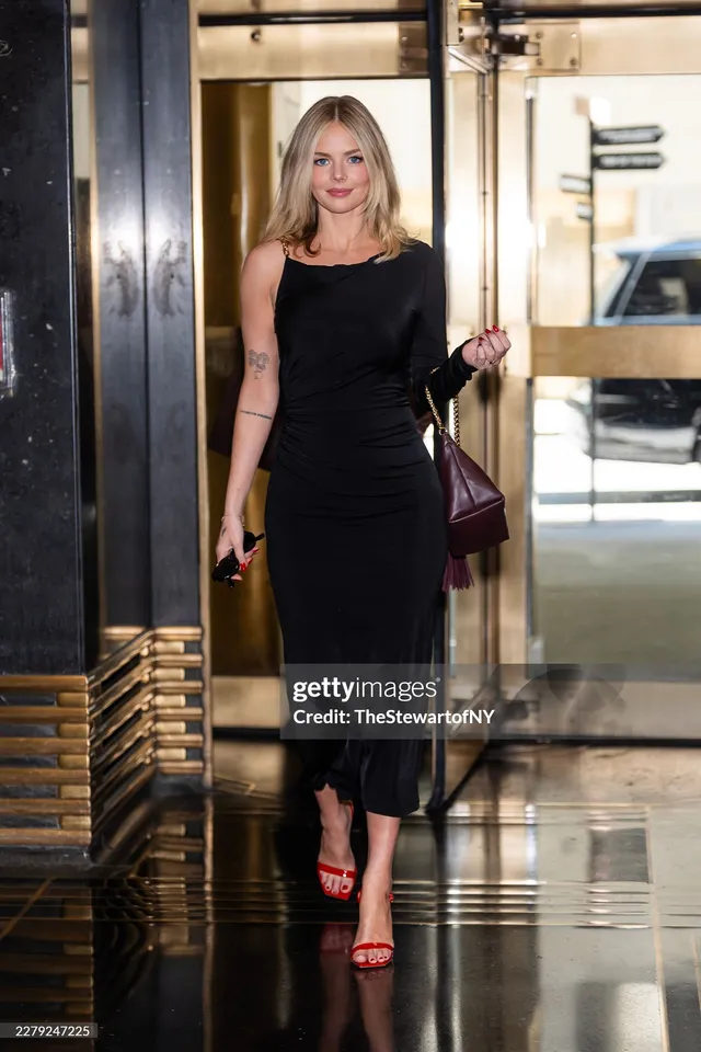 Samara Weaving standing indoors in a black dress and red heels.