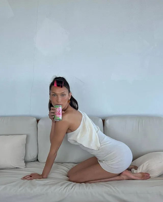 Bella Hadid kneeling barefoot on a white sofa.