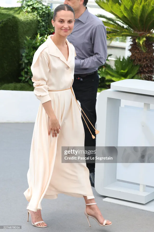 Alicia Vikander standing beside a photo call display in a cream outfit.