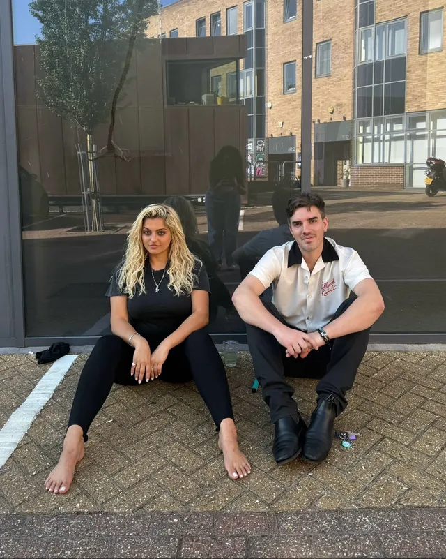 Bebe Rexha sitting barefoot on a curb beside another adult.