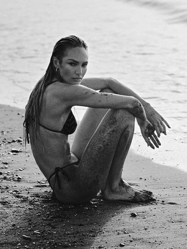 Candice Swanepoel sitting barefoot at the shoreline in a monochrome portrait.