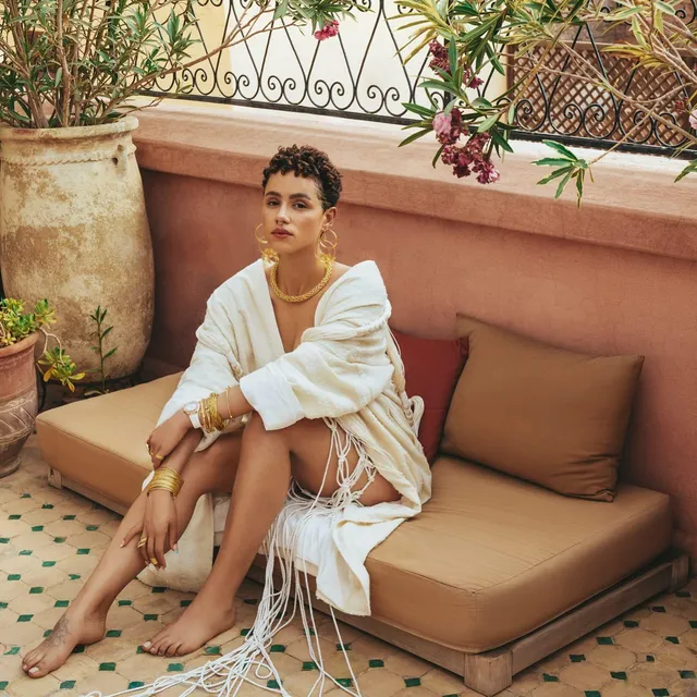 Nathalie Emmanuel sitting on an outdoor sofa in a pale outfit.