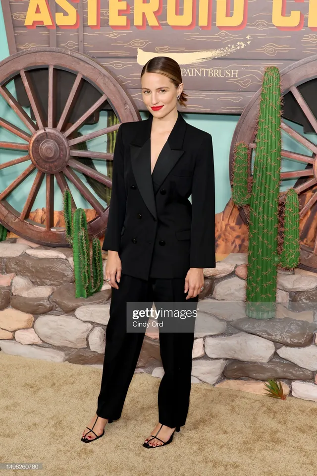 Dianna Agron standing beside a film poster in a dark tailored outfit.