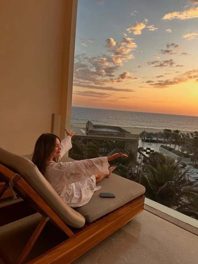 Olivia Holt reclining barefoot on an outdoor lounger in a pale shirt at sunset.