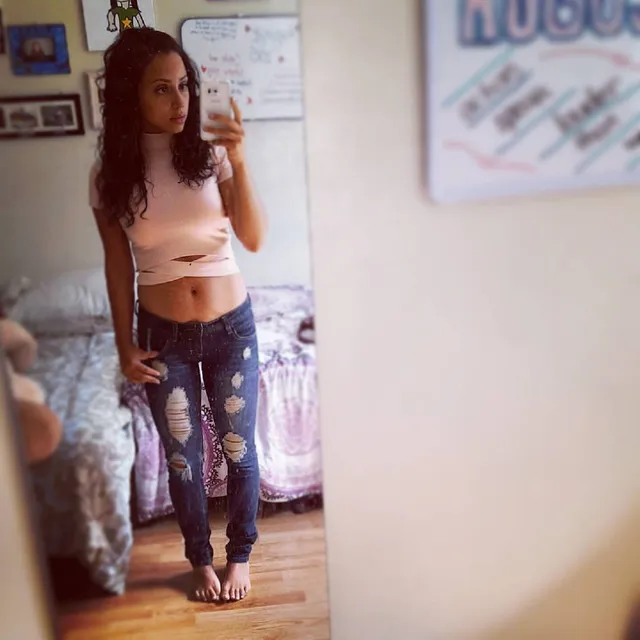 Kiana Madeira taking a mirror selfie barefoot in jeans and a pale cropped top.