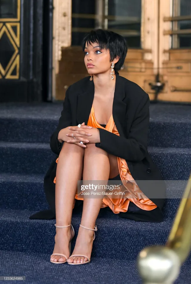 Whitney Peak sitting on steps in an orange dress, black blazer and pale heels.