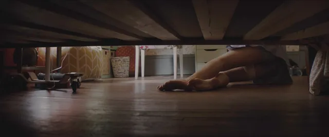 Chloë Grace Moretz lying beneath a bed with bare feet visible in a screen image.
