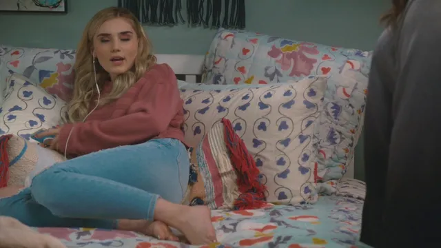 Meg Donnelly sitting barefoot on a sofa in a red top and blue trousers.