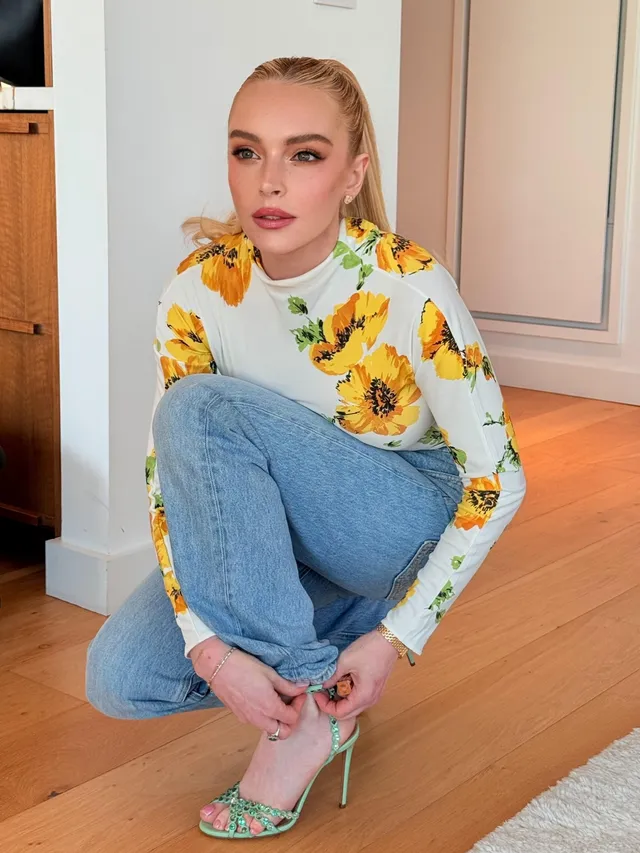 Lindsay Lohan sitting in jeans and a floral top with green heels.