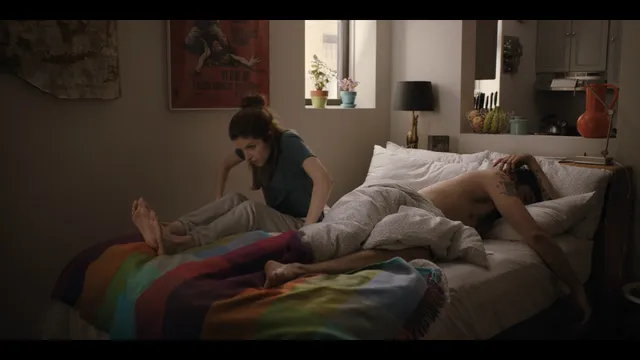 Anna Kendrick reclining barefoot on a bed beside another adult.