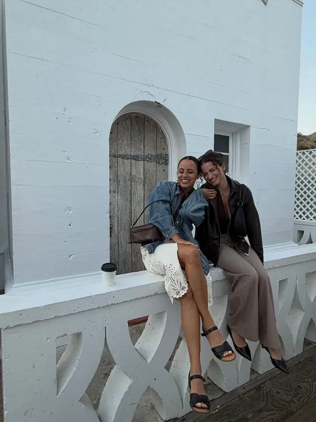 Kiana Madeira sitting beside a companion on a balcony in a pale dress and black heels.