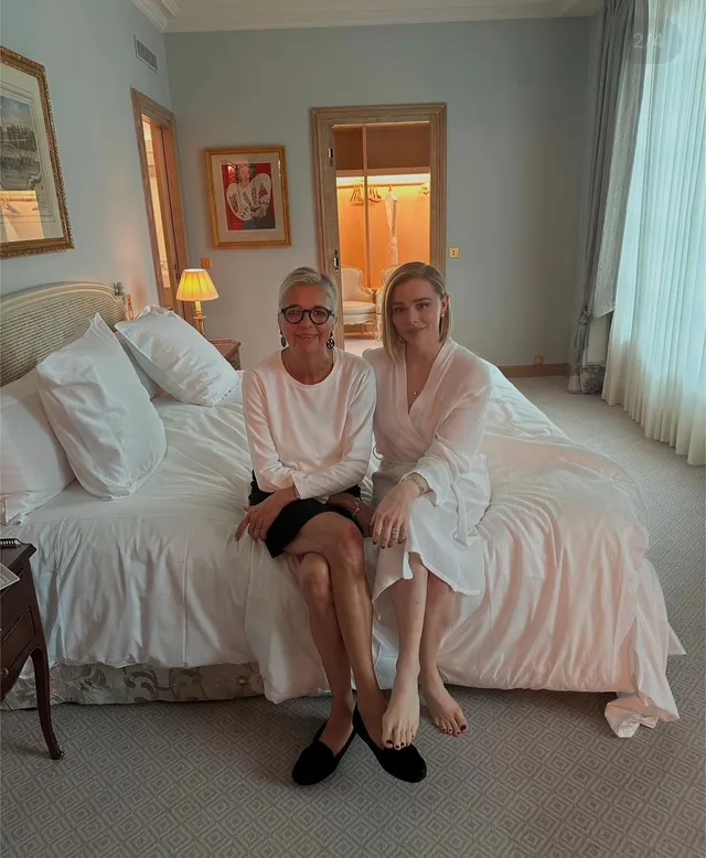 Chloë Grace Moretz sitting on a bed beside another person in a white robe and flats.