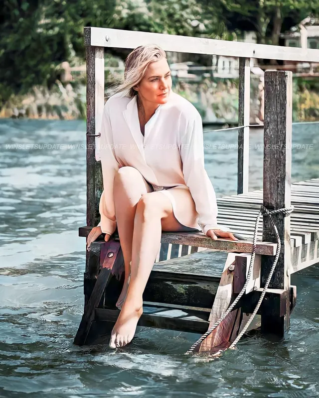 Kate Winslet sitting barefoot by the water in a light outfit.