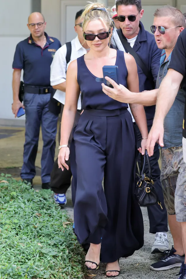 Florence Pugh standing with companions in a dark outfit and heels.