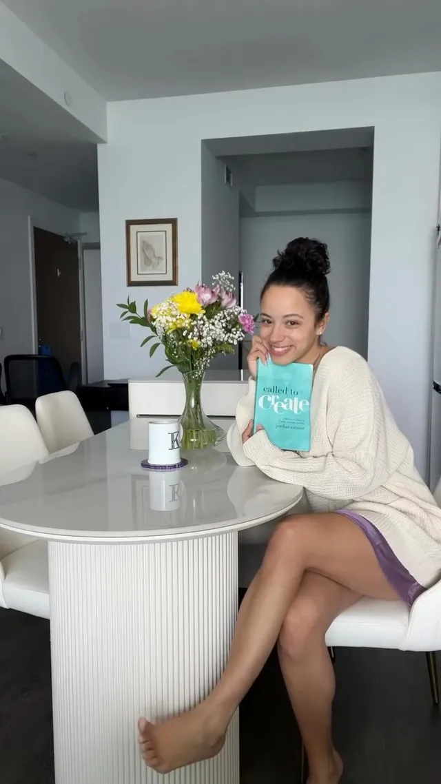 Kiana Madeira sitting barefoot at a kitchen table in a cardigan and shorts.