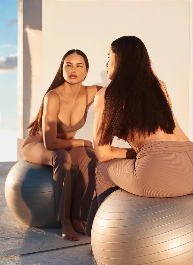 Adriana Lima sitting barefoot on an exercise ball beside a mirror.